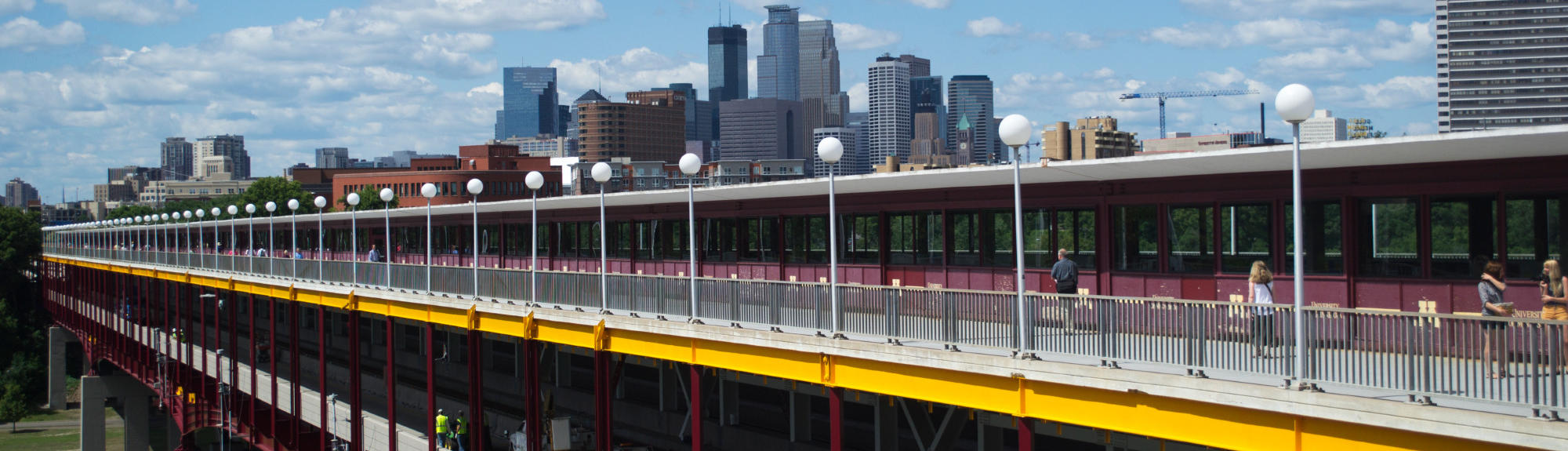 Students cross the bridge spanning East and West Bank with the Minneapolis Skyline in the background