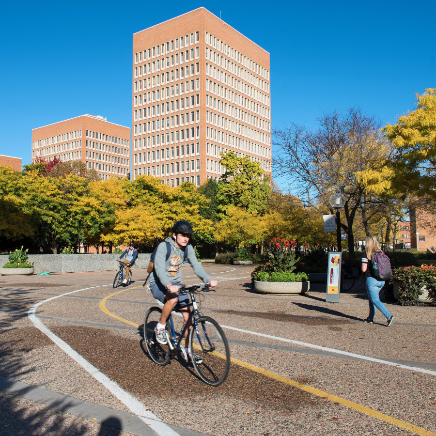 Student bikes on bike path, with other students walking, through West Bank with fall foliage in the background