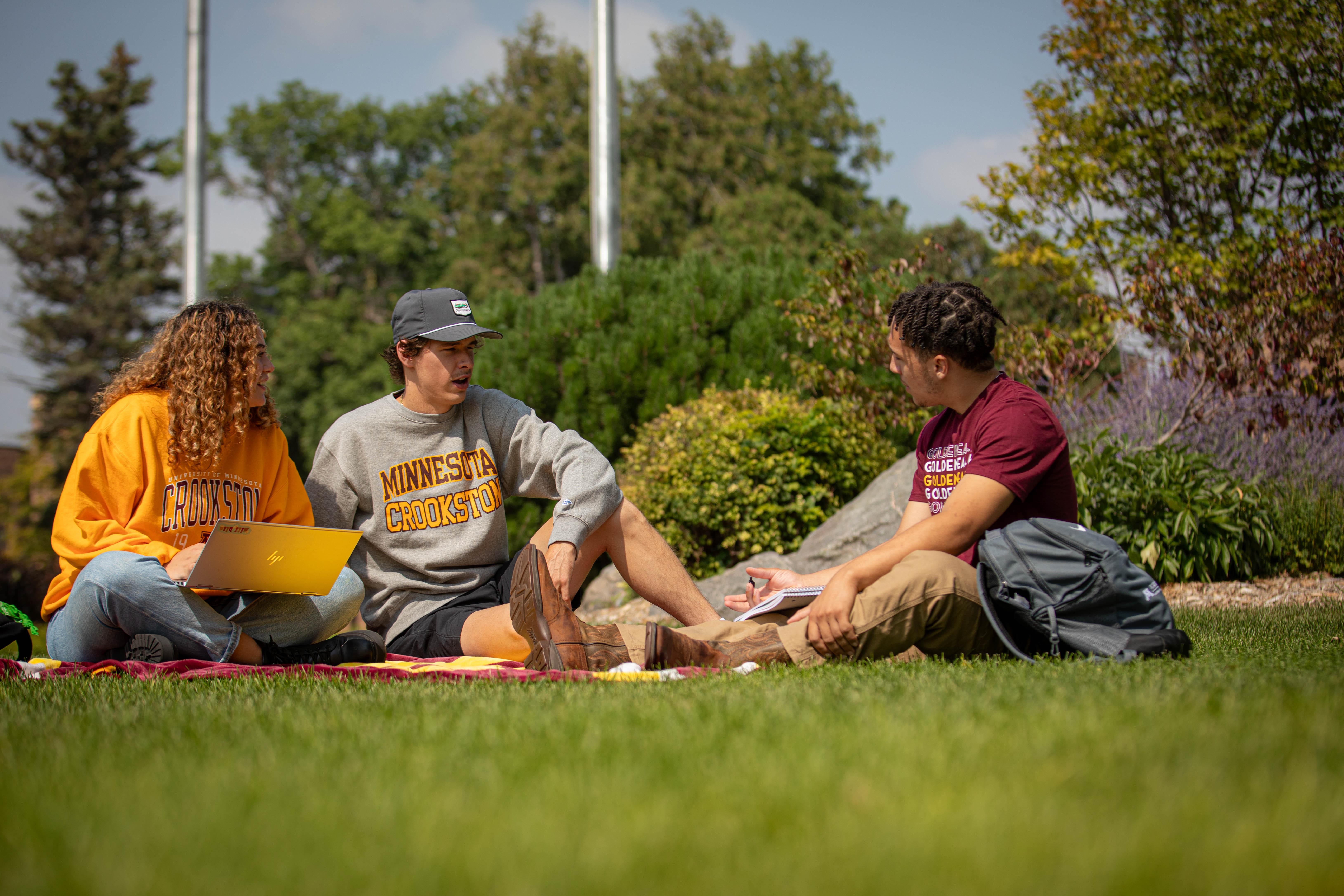 Three students talking while sitting on a blanket in the grass