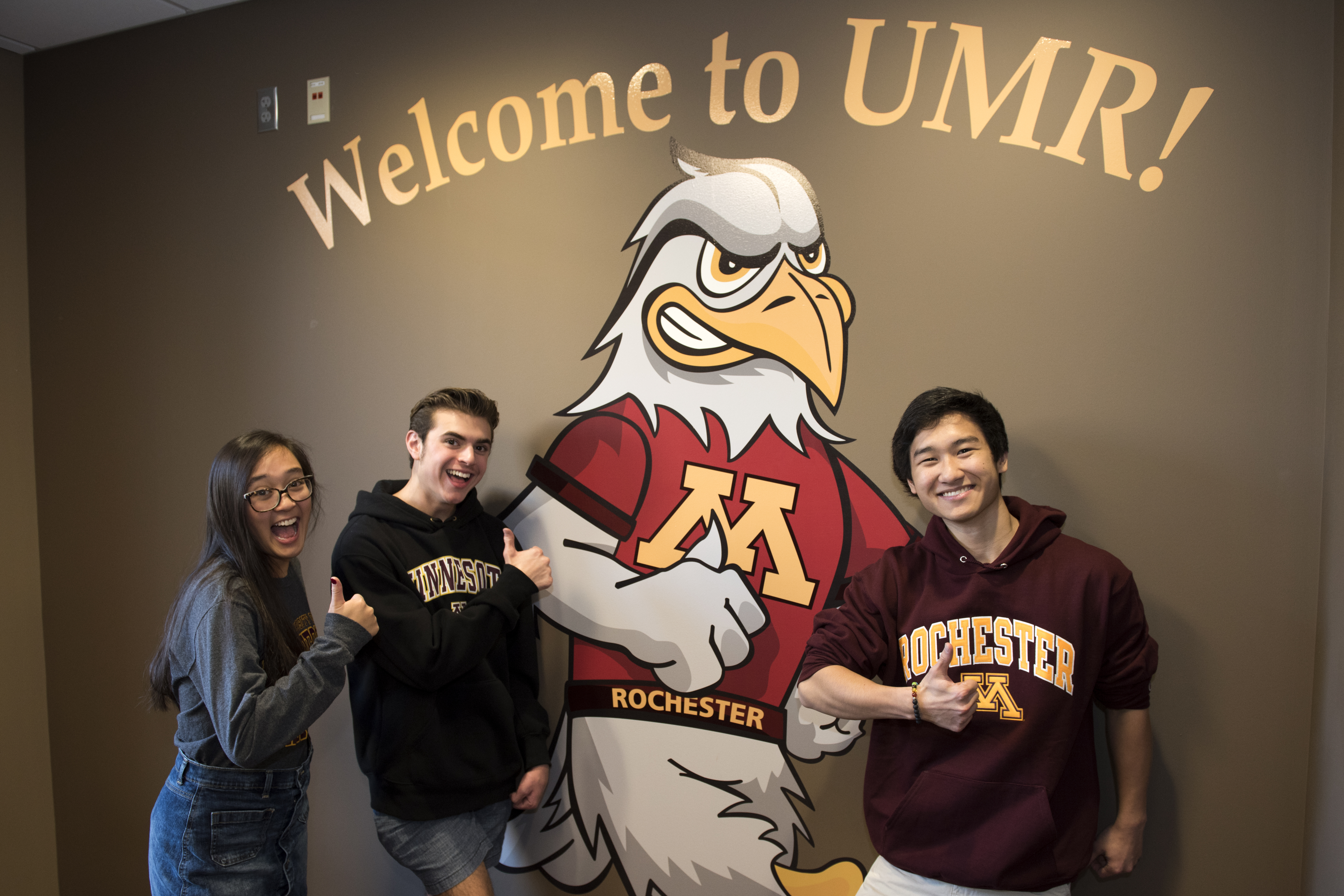 Three students smiling and giving a thumbs up, standing in front of a mural that says "Welcome to UMR!"
