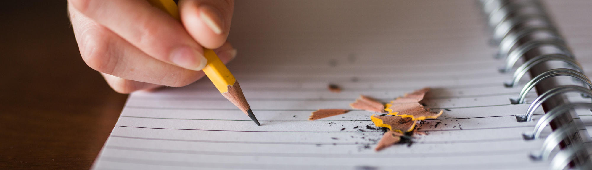Pencil shavings lay on an open notebook, while a hand writes with a pencil